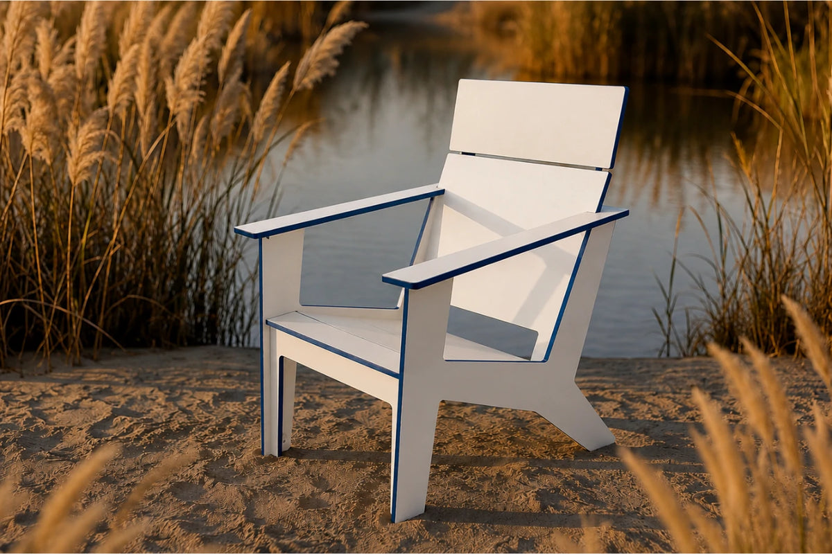 A modern white outdoor armchair with dark blue trim, positioned on a sandy bank next to a calm body of water with tall reeds in the background.
