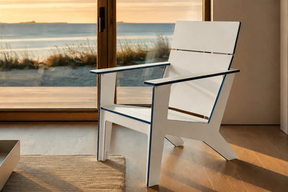 A modern white outdoor Adirondack chair with dark blue trim, positioned in front of a large glass sliding door overlooking a beach at sunset.