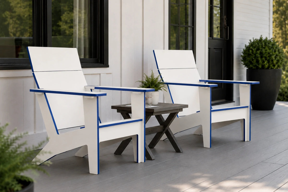 Two modern white outdoor Adirondack chairs with blue trim are shown on a gray porch next to a small dark wood side table. The background features a white house exterior with black-framed windows and a potted green shrub.