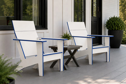 Two modern white outdoor Adirondack chairs with blue trim are shown on a gray porch next to a small dark wood side table. The background features a white house exterior with black-framed windows and a potted green shrub.