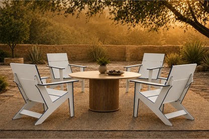 Four white outdoor Adirondack-style chairs arranged around a round slatted wood table on a patio, with trees and warm golden sunlight in the background.