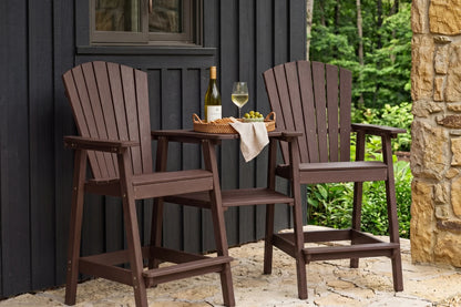 Two coffee-colored Adirondack-style outdoor bar height chairs with a matching small table between them, set on a patio in front of a black wood wall and stone column with green foliage in the background.