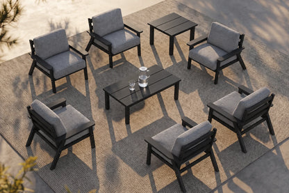 An eight-piece outdoor patio furniture set including six black-framed armchairs with gray cushions, a rectangular black coffee table, and a matching side table, arranged on a woven rug over a concrete patio.