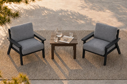 Two gray upholstered armchairs with black frames arranged around a small black side table on a woven rug, photographed in an outdoor patio setting with dappled sunlight and shadows.