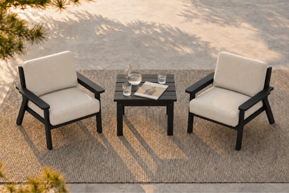 A 3-piece outdoor patio furniture set featuring two black-framed armchairs with cream cushions and a matching black slatted side table, arranged on a woven jute rug.