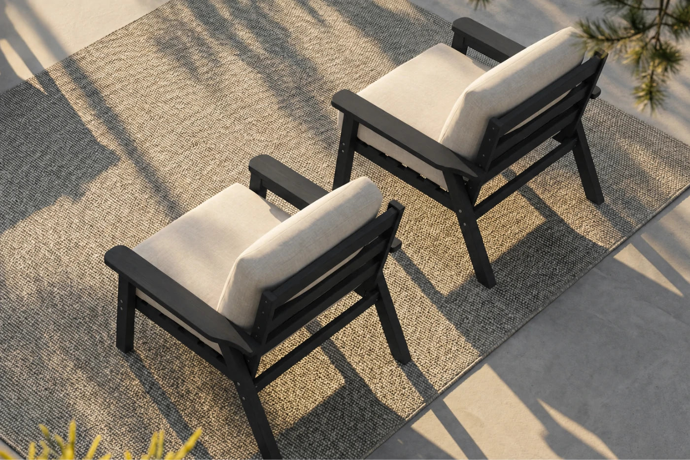 Two outdoor armchairs with black frames and creamy white cushions, arranged on a textured beige rug in a patio setting with natural shadows.