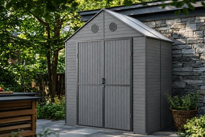 A gray resin storage shed with polycarbonate roof, shown outdoors next to a stone wall and green trees.
