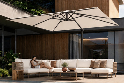 A large beige patio umbrella with a black metal frame is open over a modern outdoor sectional sofa. A woman is reclining on the white-cushioned sofa with brown accent pillows, set against a contemporary house with wood paneling and glass windows.