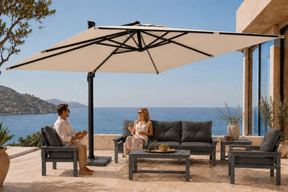 A large beige patio umbrella with a black frame is set up on a stone terrace overlooking the ocean, with a couple seated under it beside a dark gray outdoor sofa and coffee table.