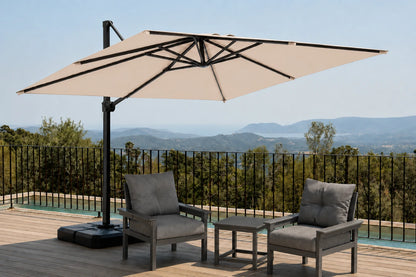 A large beige patio umbrella with a black cantilever pole and base, shown on a wooden deck with two gray outdoor armchairs and a small side table, overlooking a scenic mountain landscape.