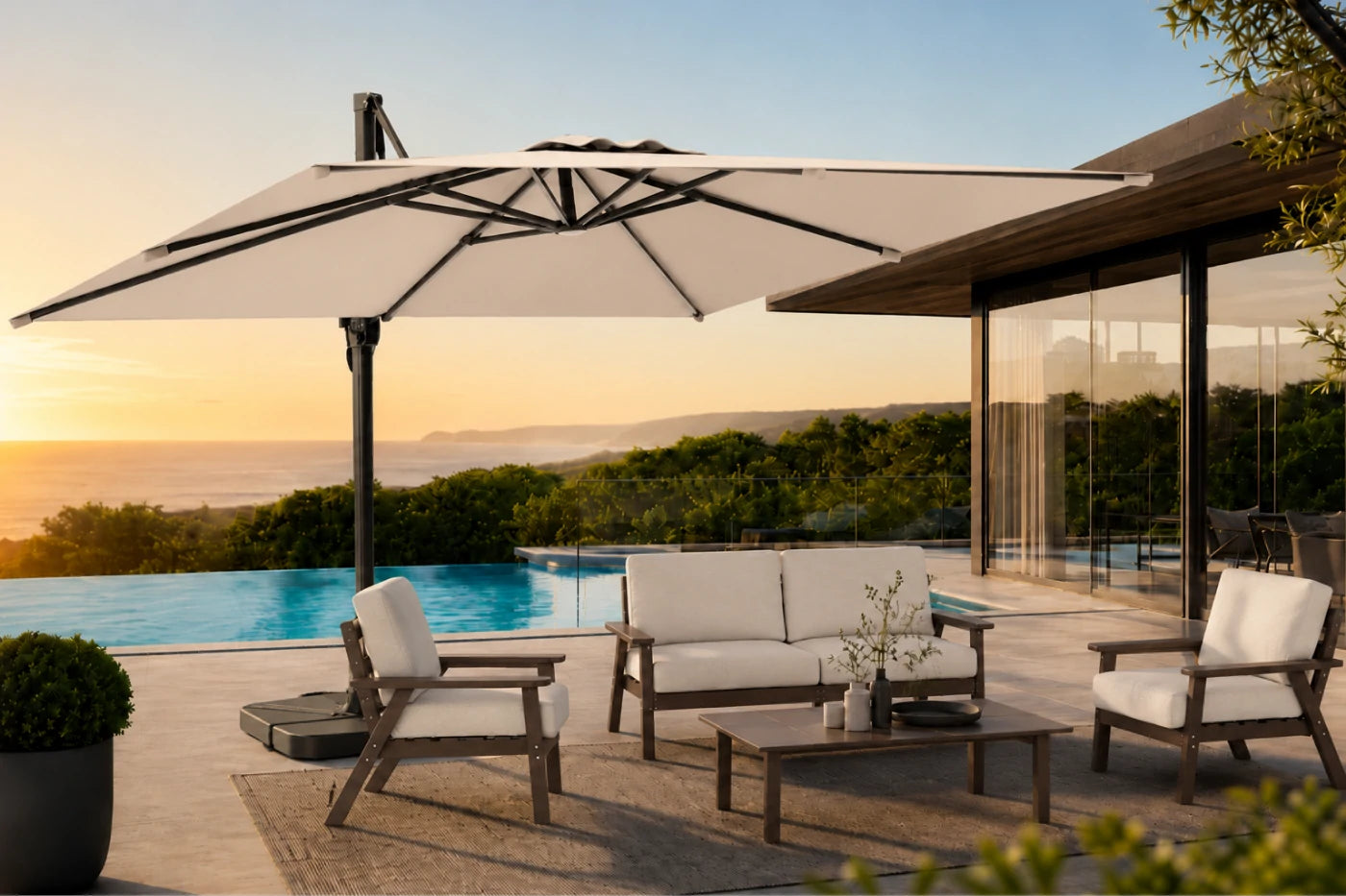 A large patio umbrella with a beige fabric canopy and black frame is shown on a luxury outdoor deck next to a swimming pool at sunset. The scene includes a wooden sofa, two matching armchairs, and a coffee table, with a modern house and ocean view in the background.
