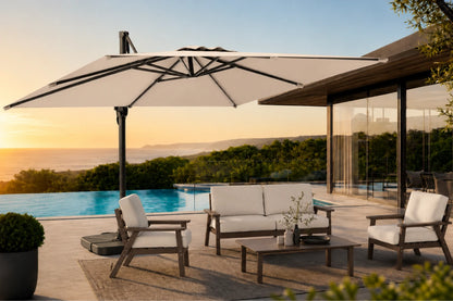A large patio umbrella with a beige fabric canopy and black frame is shown on a luxury outdoor deck next to a swimming pool at sunset. The scene includes a wooden sofa, two matching armchairs, and a coffee table, with a modern house and ocean view in the background.