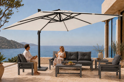A large cream-colored patio umbrella with a black frame is set up on a stone terrace, shading a modern outdoor seating area with a sofa, armchairs, and a coffee table. A man and a woman are seated under the umbrella, with a blue sea and hills in the background.