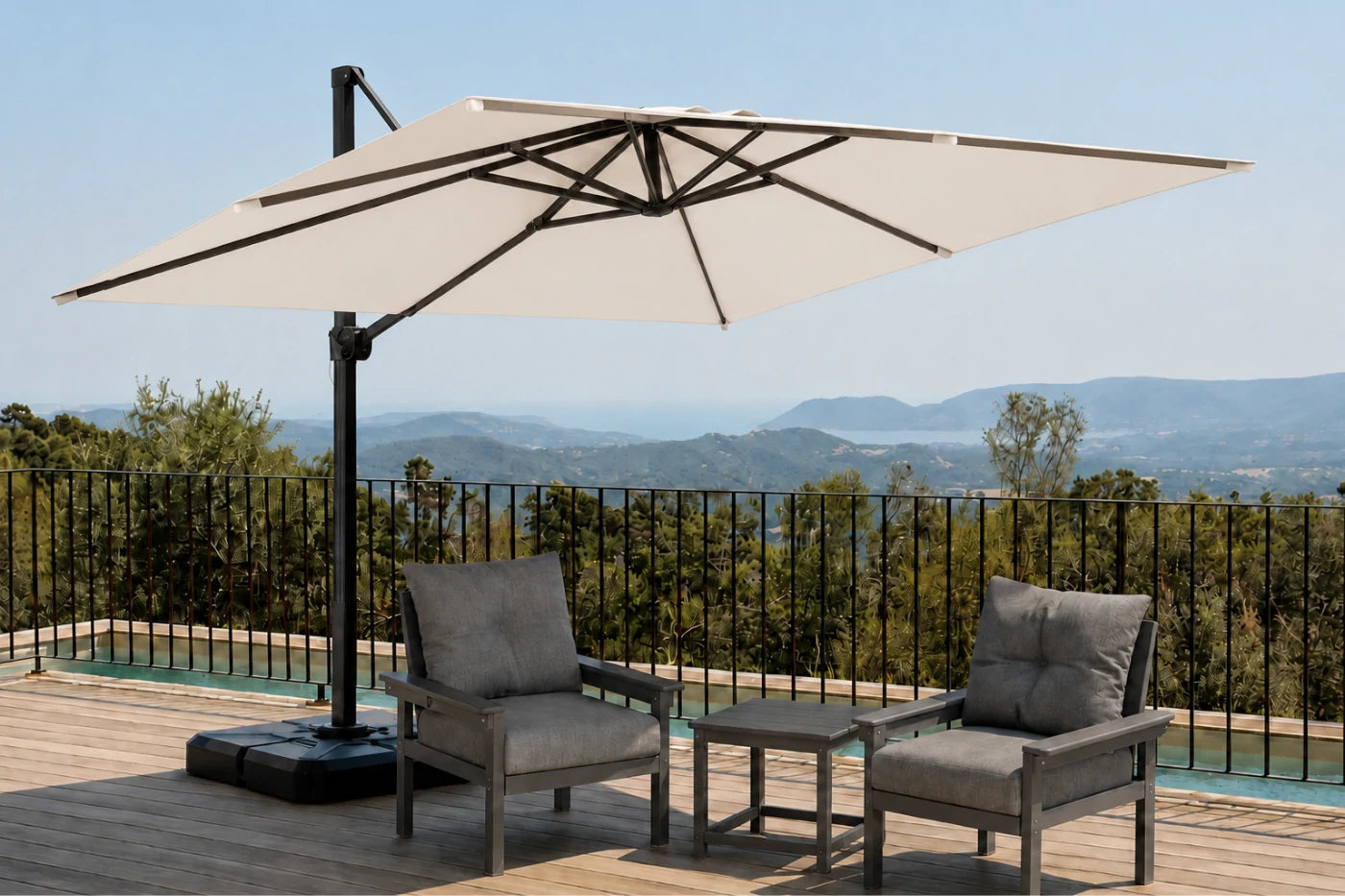 A large creamy white patio umbrella with a black cantilever pole stands on a wooden deck beside a pool, shading two gray outdoor armchairs and a small side table. A black metal railing overlooks a scenic mountain landscape under a clear blue sky.