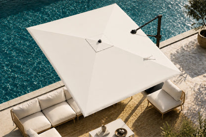 A large white patio umbrella with a black metal arm is positioned over a sunken outdoor seating area next to a swimming pool. The patio features light beige stone flooring, a white sectional sofa, and a matching armchair.