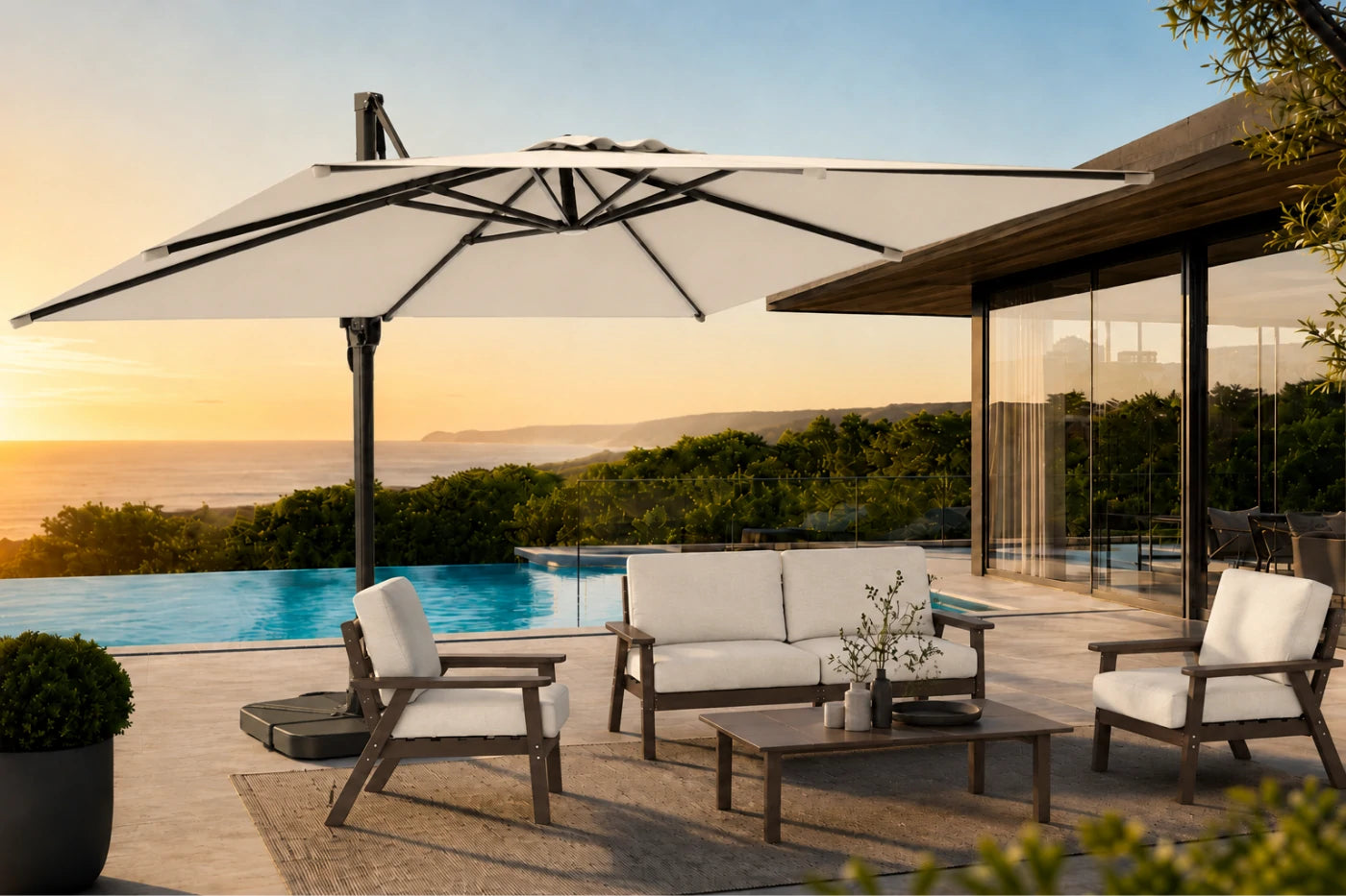 Outdoor patio furniture set with a large white cantilever umbrella, a white sofa, two matching armchairs, and a coffee table on a stone deck beside a pool at sunset.