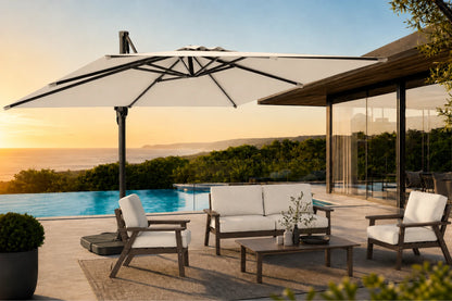 Outdoor patio furniture set with a large white cantilever umbrella, a white sofa, two matching armchairs, and a coffee table on a stone deck beside a pool at sunset.