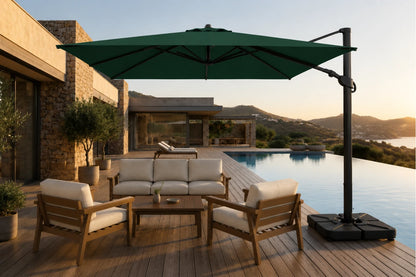 A large dark green cantilever patio umbrella with a black pole and weighted base, shading a wooden outdoor sofa and two matching armchairs on a poolside deck at sunset.