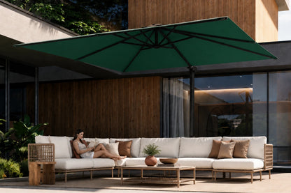 A modern outdoor patio scene with a large dark green patio umbrella shading a white sectional sofa with brown accent pillows. A woman is reclining on the sofa, and the setting includes a wooden wall, glass windows, and a coffee table.