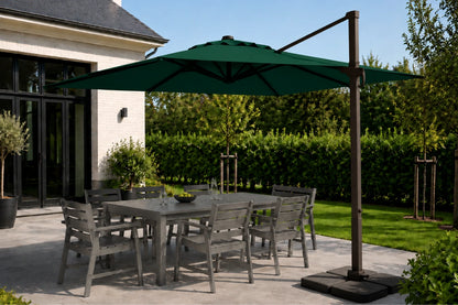 A dark green cantilever patio umbrella with a square base and brown metal pole, shading a gray outdoor dining table and chairs on a patio next to a house.