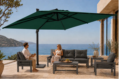 A large dark green patio umbrella shades an outdoor seating area on a sunlit terrace overlooking the ocean. Two people are seated under the umbrella on gray patio furniture beside a modern building.