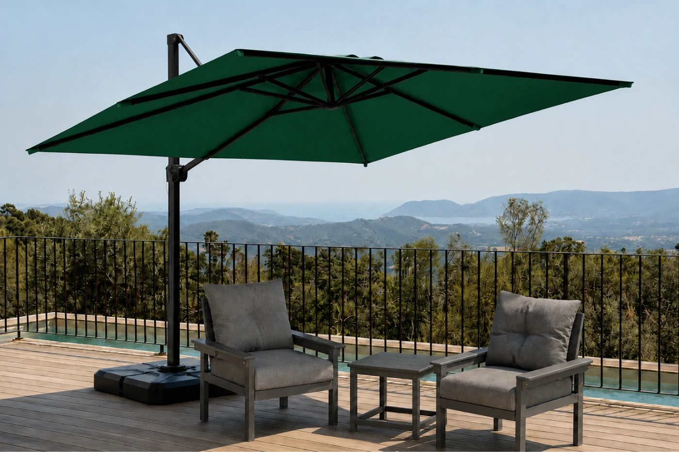 A large dark green cantilever patio umbrella with a black metal frame and base, shading two gray outdoor lounge chairs and a small side table on a wooden deck. A black metal railing and a scenic mountain landscape are visible in the background.
