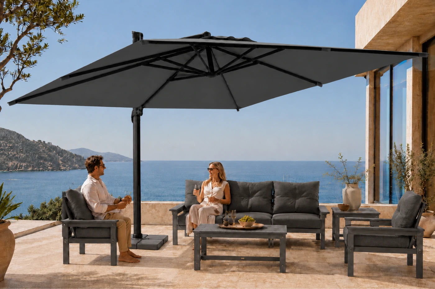 A large dark gray cantilever patio umbrella is shown on a stone terrace overlooking the sea, with two people seated under and around the umbrella and outdoor furniture.