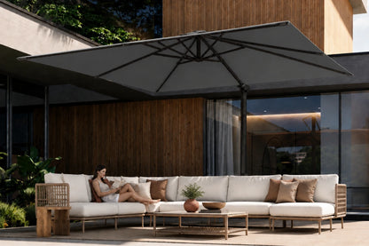 A modern outdoor patio scene with a large dark gray patio umbrella over a long white sectional sofa with brown accent pillows. A woman is reclining on the sofa, and the setting includes a wooden wall, glass windows, and a coffee table in the foreground.