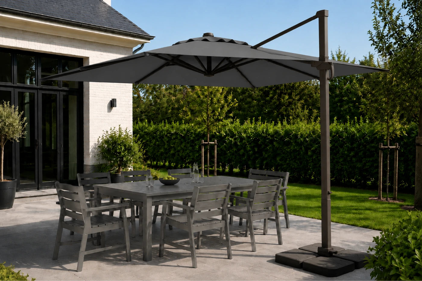A large dark gray cantilever patio umbrella with a square base is shown outdoors on a patio next to a dining set. The setting includes a modern house, green hedges, and a lawn under a clear blue sky.