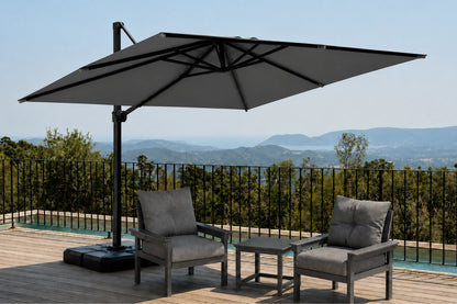 A large dark gray cantilever patio umbrella with a square gray canopy is shown on a wooden deck beside a pool, with two gray lounge chairs and a table in front. A real outdoor setting with a railing and distant hills is visible, so this is Lifestyle rather than Product/Packaging.