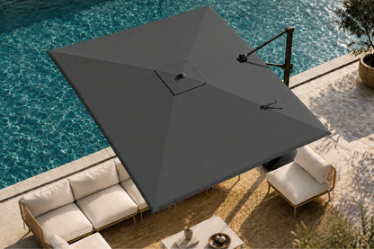 A large cantilever patio umbrella in dark gray shading an outdoor lounge area next to a swimming pool. The umbrella features a black metal support arm and is shown from an elevated angle over a beige stone patio with cream-colored outdoor furniture.