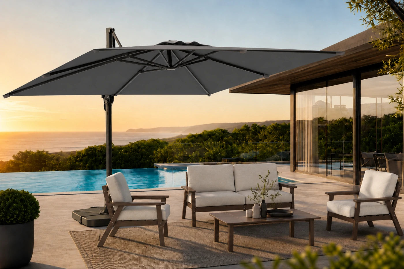 A large dark gray cantilever patio umbrella is shown in a luxurious outdoor setting next to a swimming pool and modern house at sunset. The scene includes a wooden three-seater sofa, two matching armchairs with white cushions, and a low wooden coffee table on a neutral rug.