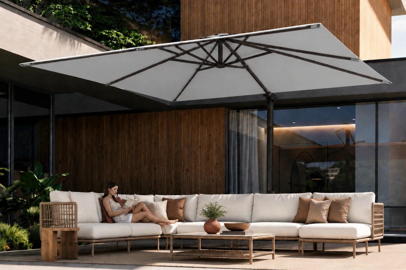 A large square patio umbrella in a gray fabric with a black metal frame, shading a modern outdoor sectional sofa with white cushions and brown pillows. A woman is reclining on the sofa, and the setting is a contemporary house with wood paneling and glass windows.