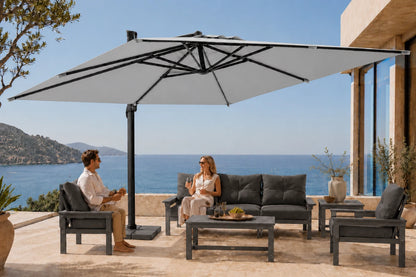 A large patio umbrella with a black frame and gray canopy is shown outdoors on a terrace, shading a modern outdoor seating area with a sofa, two armchairs, and a coffee table. A man and woman are seated under the umbrella, with a blue ocean and hills in the background.