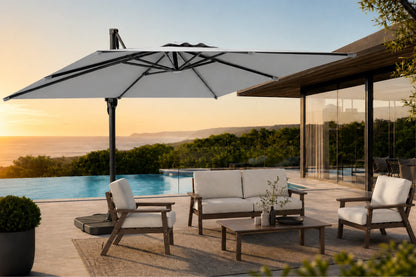 A large cantilever patio umbrella in gray with a black frame is shown outdoors next to a pool and modern house at sunset. The scene includes a white sofa, two matching armchairs, and a coffee table on a patio.