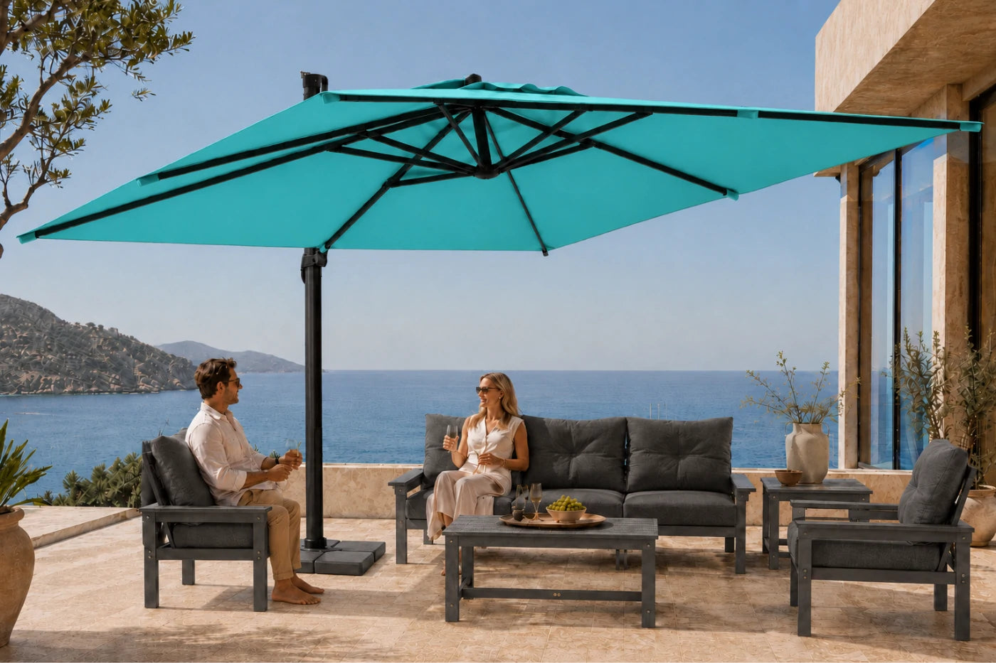 A large turquoise cantilever patio umbrella with a black frame is shown outdoors on a stone terrace beside the sea, with two people seated under it on a dark gray outdoor sofa and chairs.