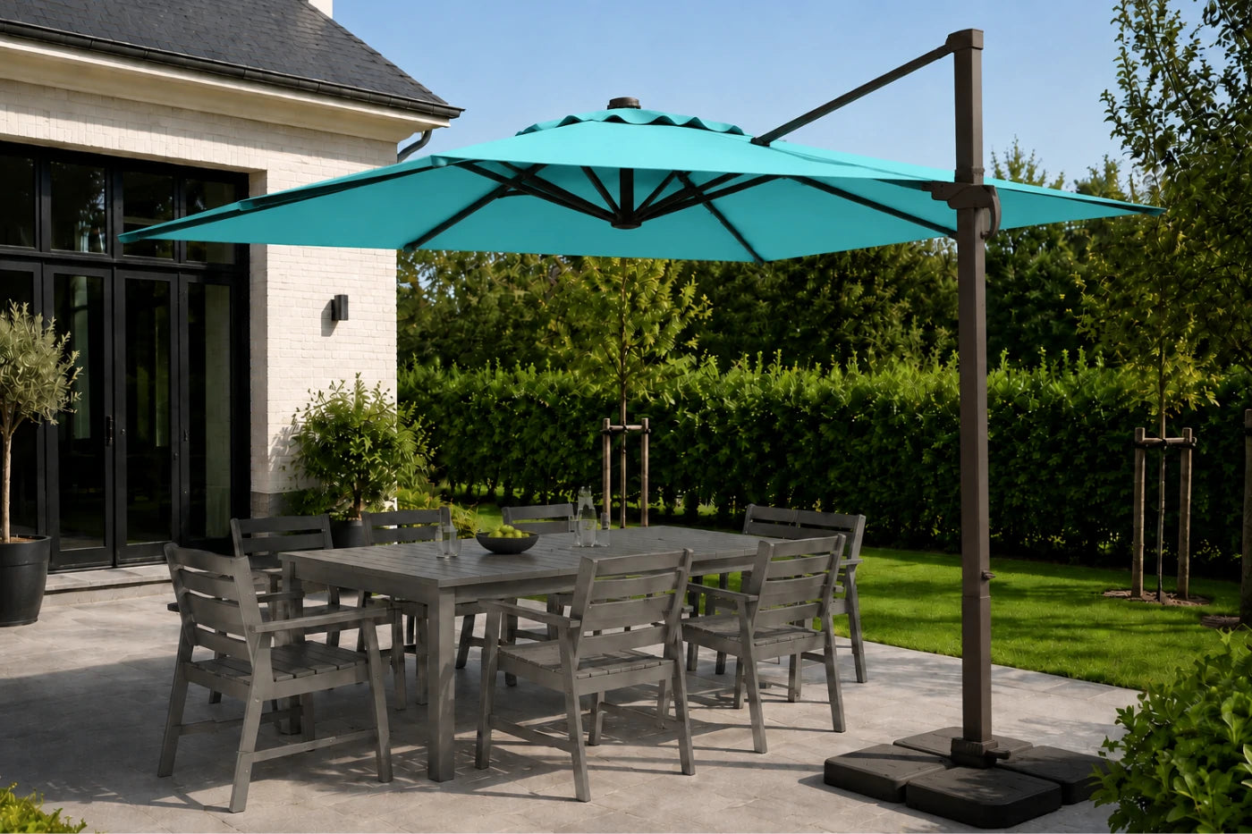 A large turquoise cantilever patio umbrella with a dark metal pole and base, shading a gray outdoor dining table and chairs on a patio next to a house.