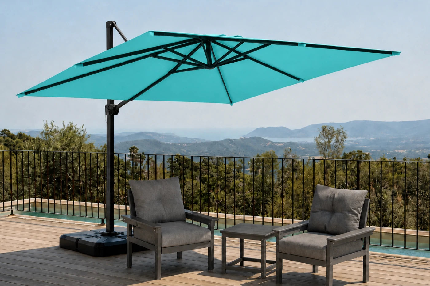 A large teal cantilever patio umbrella with a black pole and base, shown on a wooden deck beside two gray outdoor armchairs and a small side table, with a scenic mountain landscape in the background.