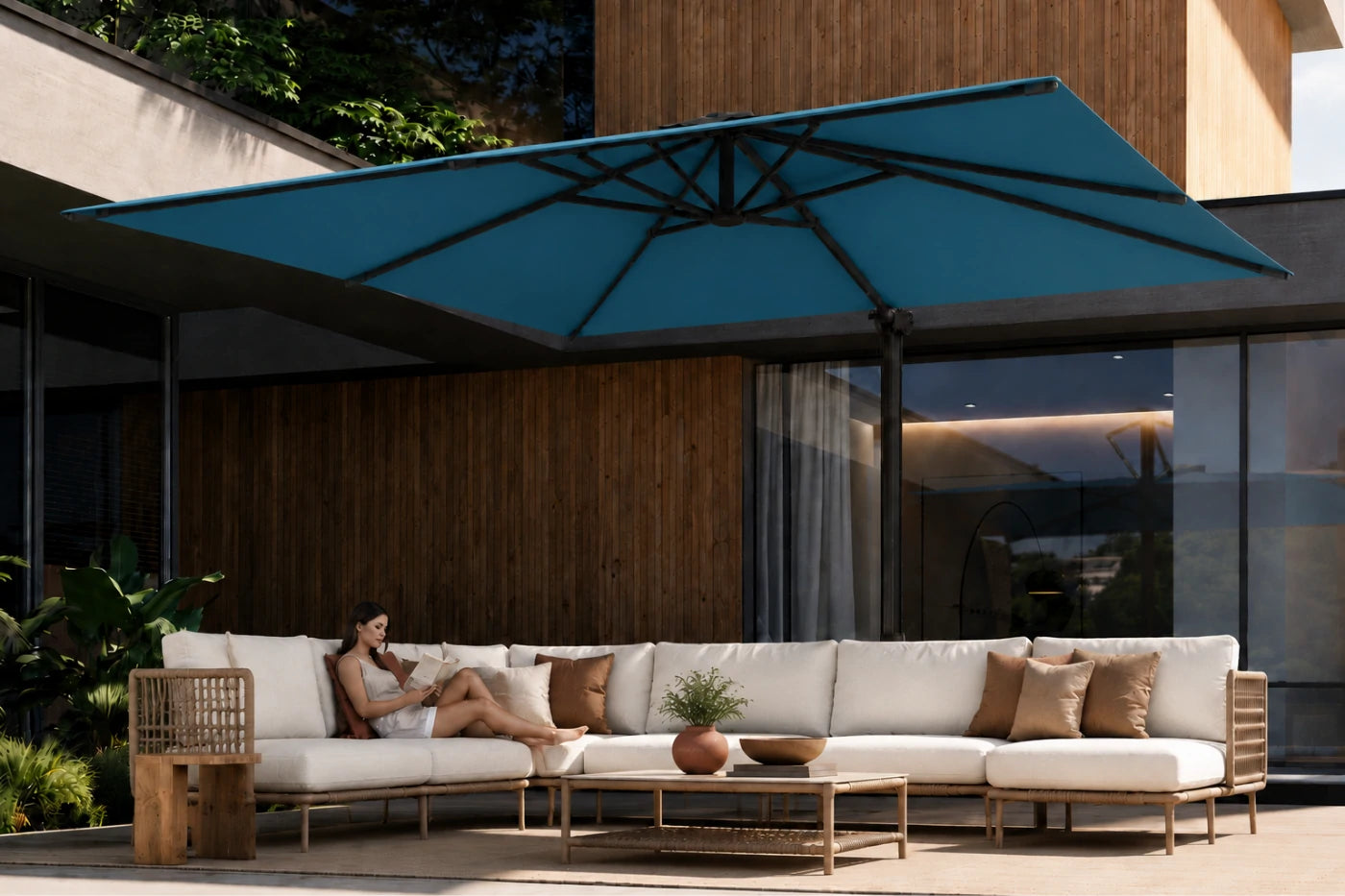 A large lake blue patio umbrella shades an outdoor lounge area with a white sectional sofa, wooden coffee table, and a woman reclining on the sofa in front of a modern building with wood siding.