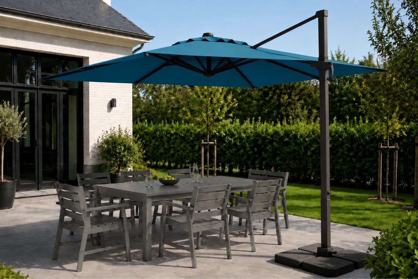 A large lake blue cantilever patio umbrella with a dark metal pole and base, shading a gray outdoor dining table and chairs on a paved patio next to a house.