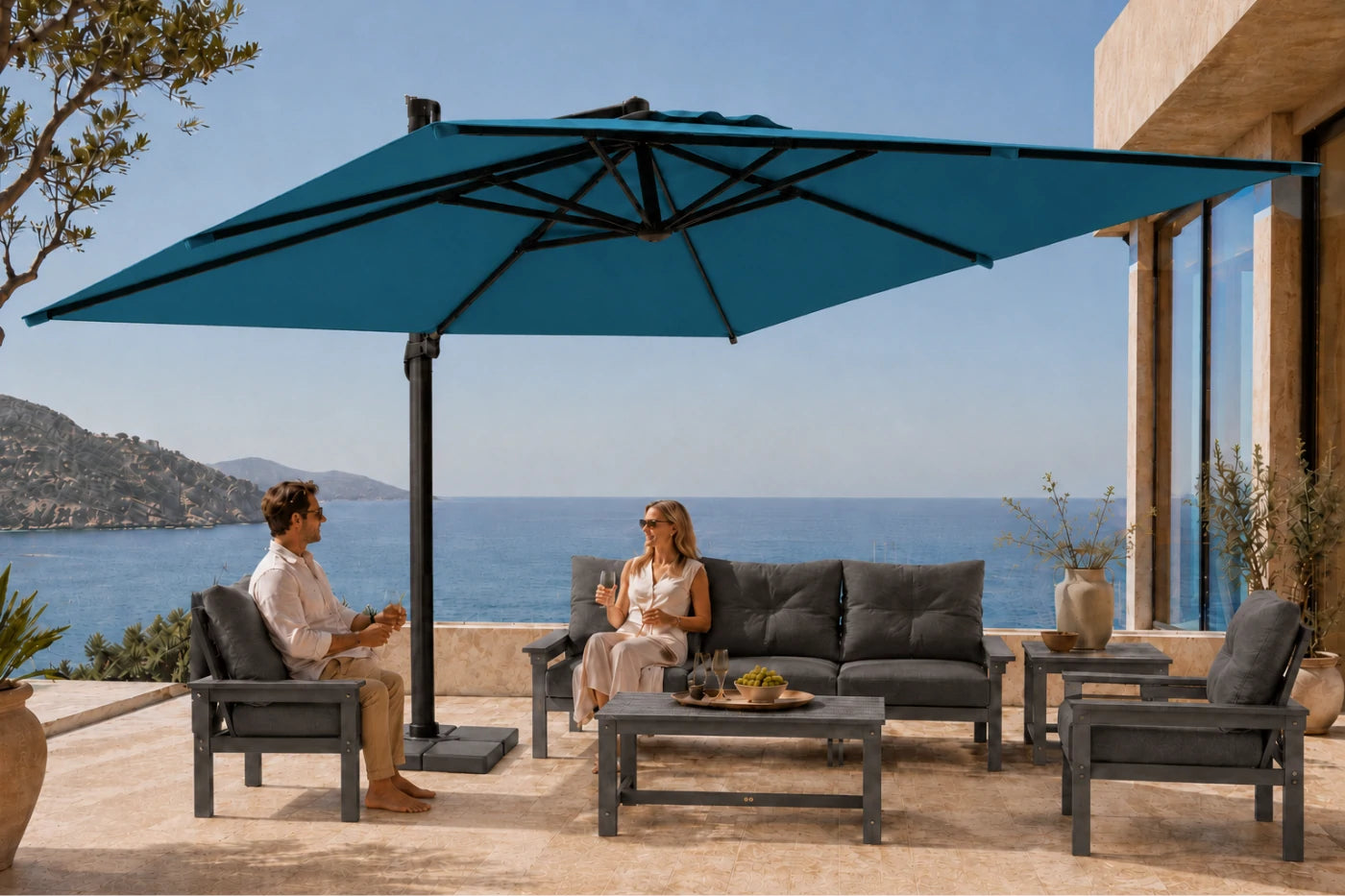 A large lake blue cantilever patio umbrella shades an outdoor lounge set with dark gray cushions, tables, and chairs on a sunlit terrace overlooking the sea. A man and woman are seated under the umbrella, conversing in a luxurious setting.