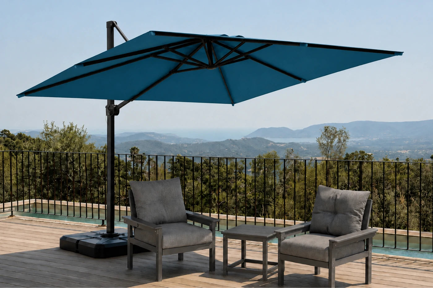 A large lake blue cantilever patio umbrella with a black metal frame and base, set on a wooden deck next to a pool with two gray outdoor armchairs and a small side table. A scenic mountain landscape is visible in the background under a clear blue sky.