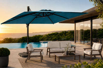 A large lake blue patio umbrella shading a modern outdoor seating area with a pool at sunset. The scene includes a sofa, two armchairs, a coffee table, and a house with glass walls in the background.