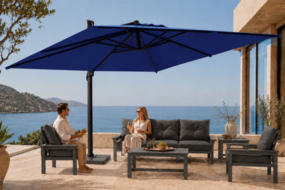 A large navy blue patio umbrella with a black pole and base is set up on a stone terrace overlooking the ocean, with a man and woman sitting on dark gray outdoor furniture under it.