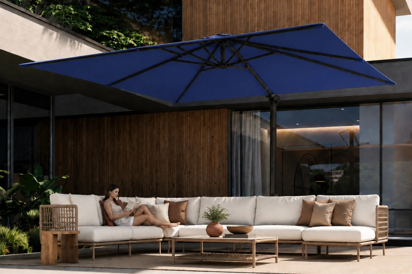 A large navy blue patio umbrella shades an outdoor sectional sofa with a woman reclining on the cushions, set against a modern building with wood paneling and glass windows.