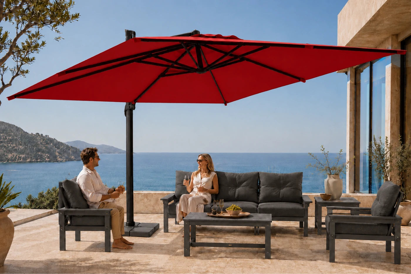 Outdoor patio furniture set with a large red cantilever umbrella, dark gray sofa, matching armchair, and coffee table on a stone terrace overlooking the ocean, with two people seated under the umbrella.