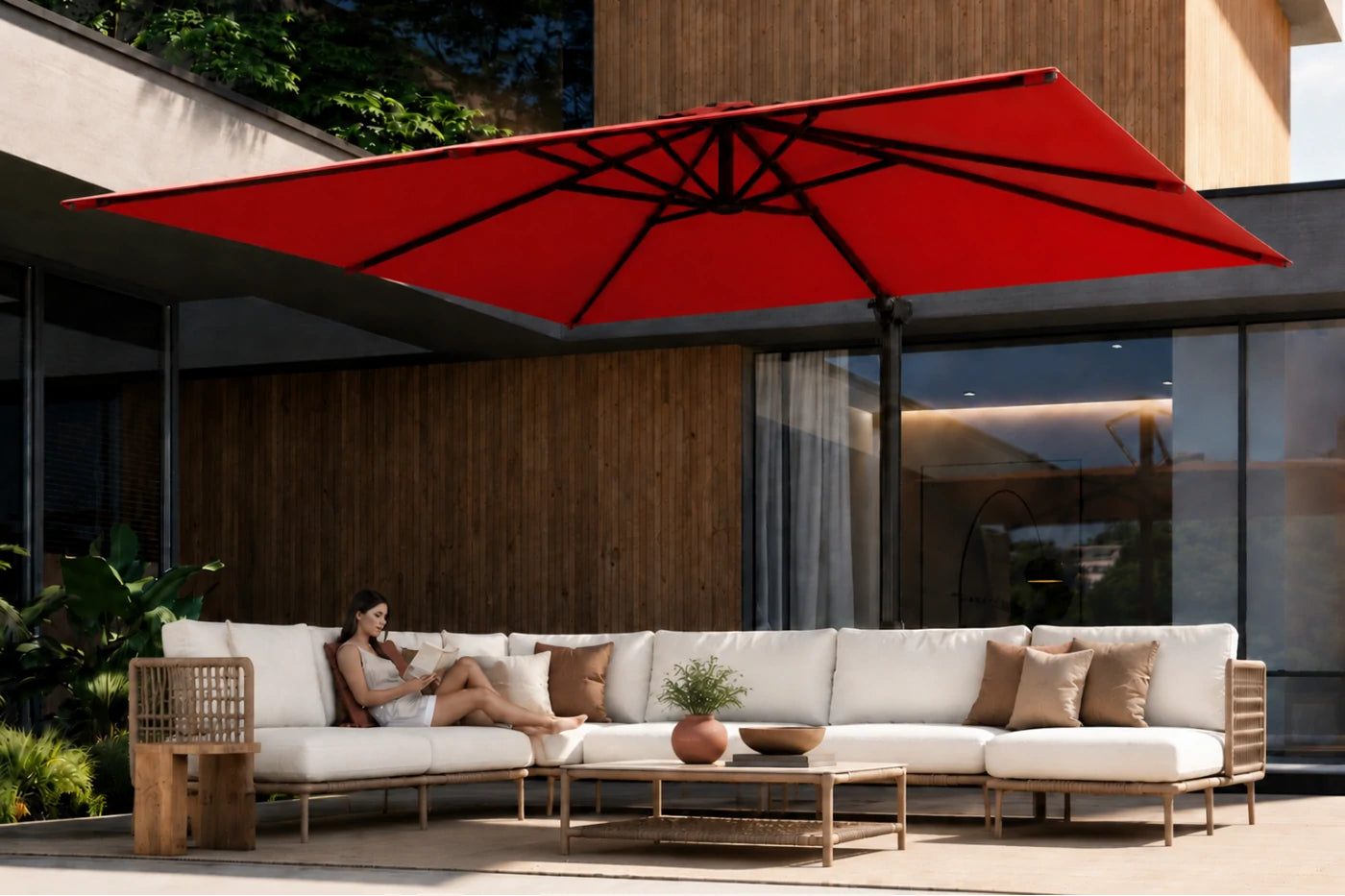 A large red patio umbrella shades an outdoor lounge area with a white sectional sofa, wooden coffee table, and a woman sitting on the sofa in front of a modern house with wood paneling.