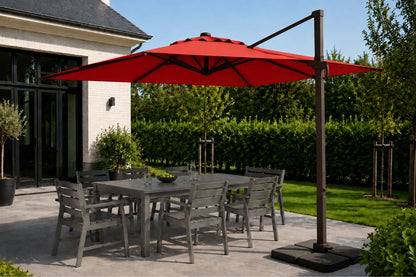 A large red cantilever patio umbrella with a dark metal frame stands over a gray outdoor dining table and chairs on a paved patio. The scene is set in a backyard with green grass, hedges, and trees under a clear blue sky.
