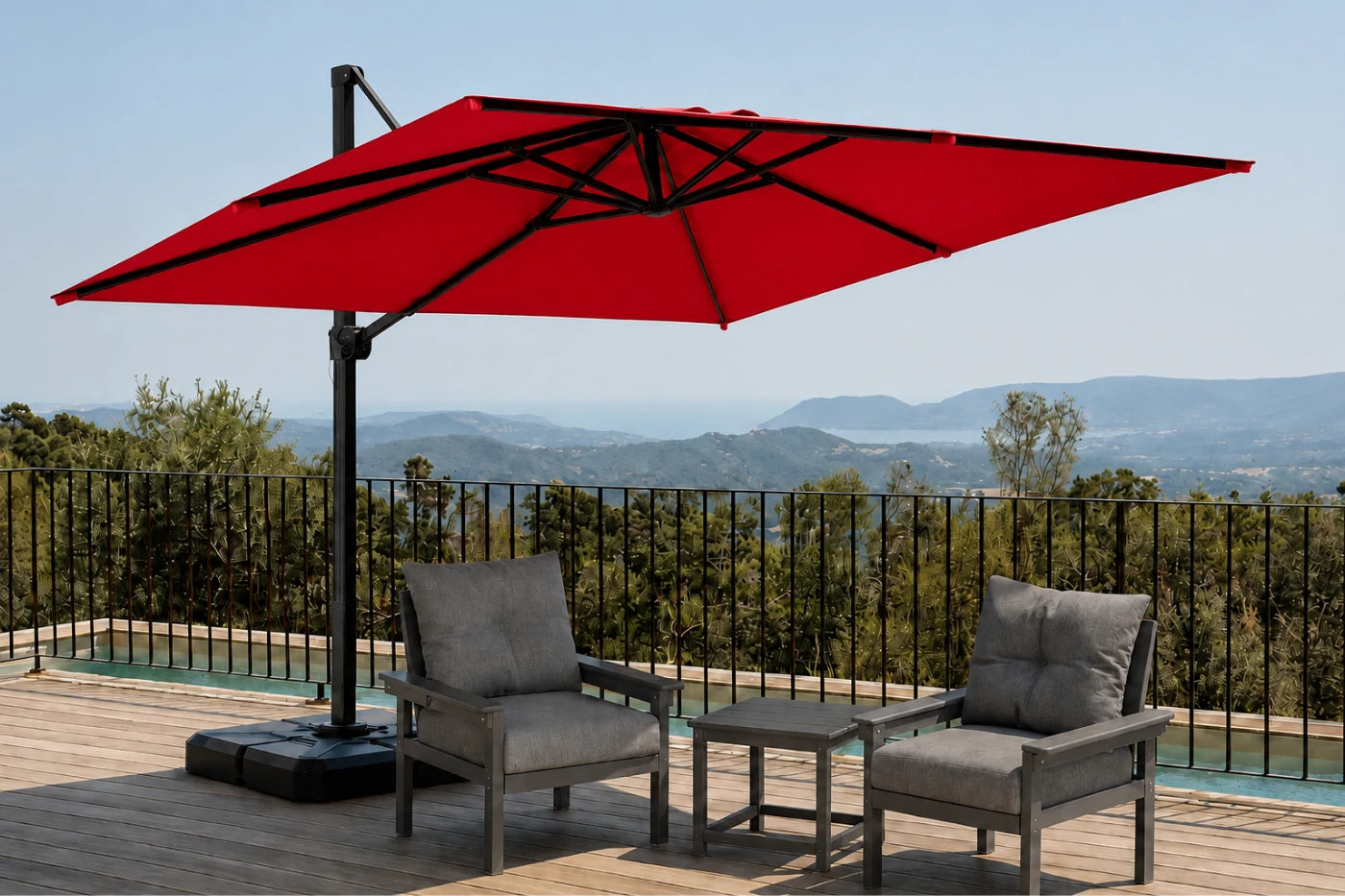 A large red cantilever patio umbrella with a black pole and base, shown on a wooden deck with two gray lounge chairs and a small side table. A black metal railing and distant green hills under a blue sky are visible in the background.